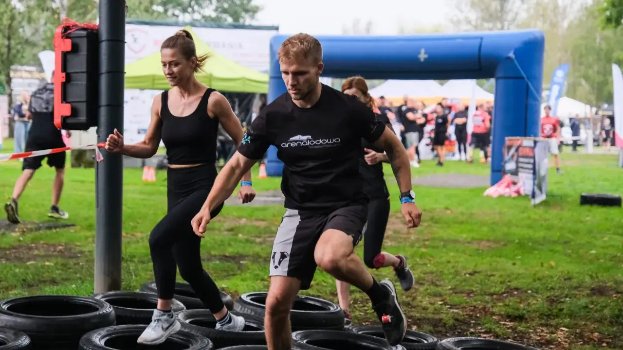 Zawodnicy pokonują tor przeszkód z opon. Kobieta i mężczyzna biegną, a za nimi inni uczestnicy. To prawdziwa survival race!