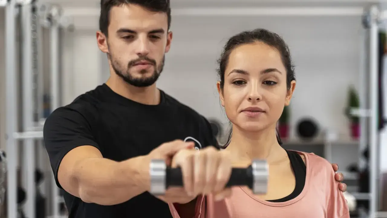 Trening anaerobowy z trenerem. Kobieta ćwiczy z hantlem, mężczyzna koryguje jej ruchy.