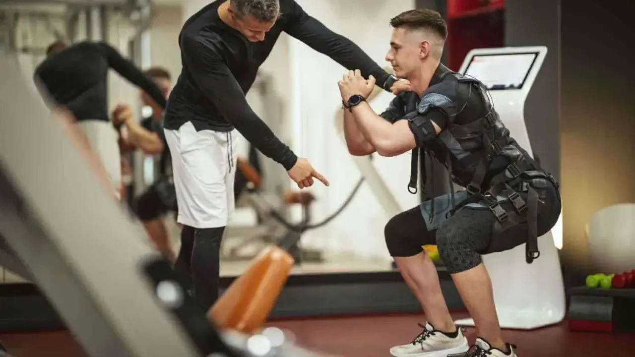 Trening EMS: Instruktor koryguje pozycję mężczyzny w kamizelce stymulującej mięśnie podczas przysiadu.