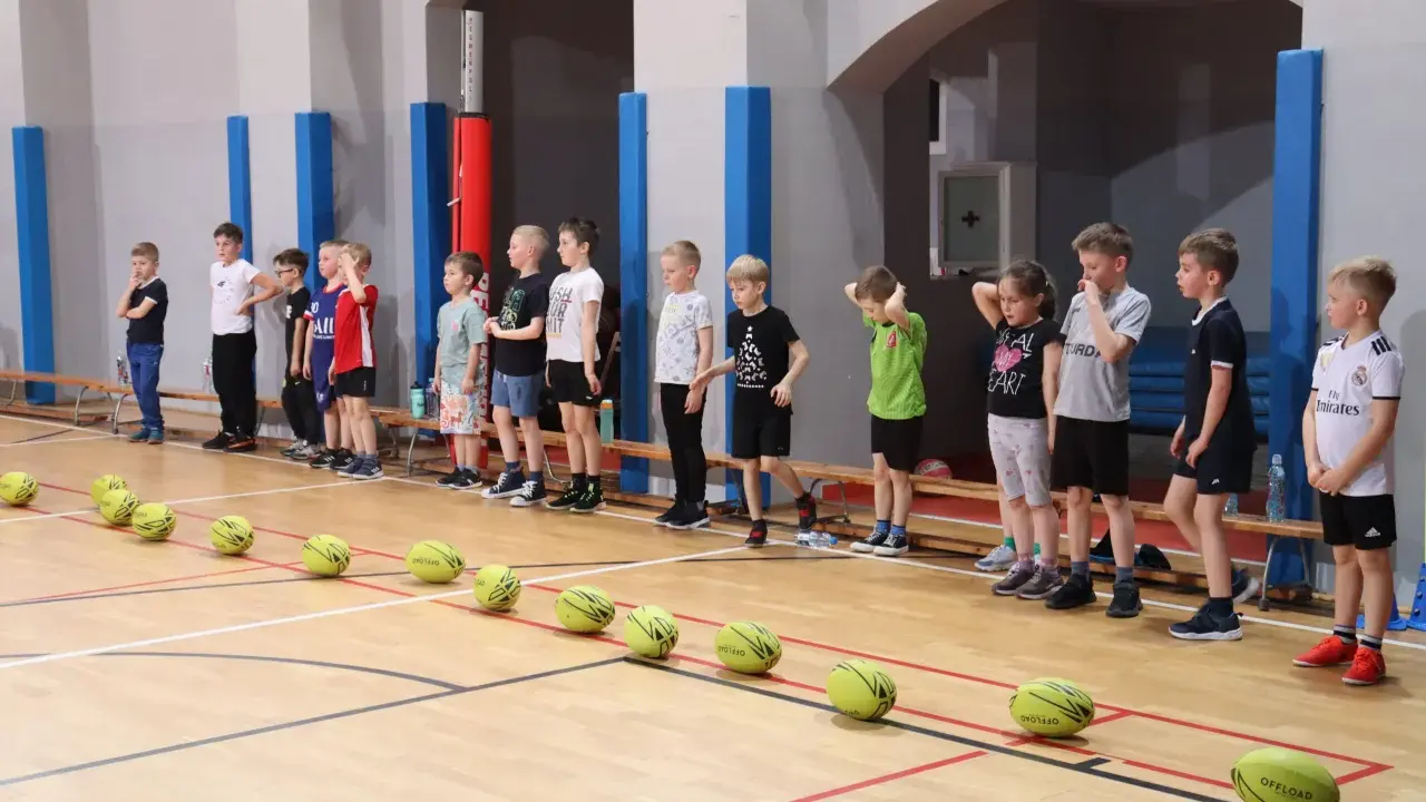 Dzieci w hali sportowej czekają na rozpoczęcie gry i zabawy. Na parkiecie leżą żółte piłki.