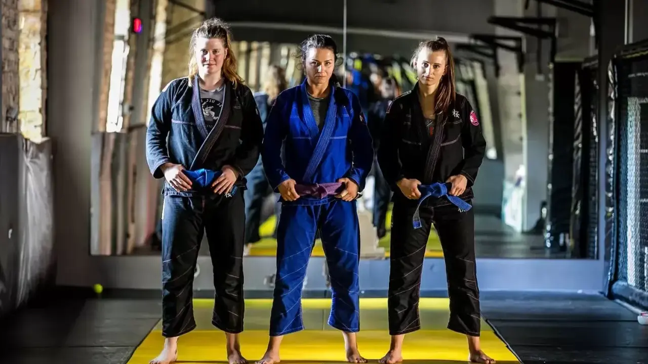 Trzy kobiety w kimonach do BJJ. Środkowa ma purpurowy pas bjj, a pozostałe niebieskie.