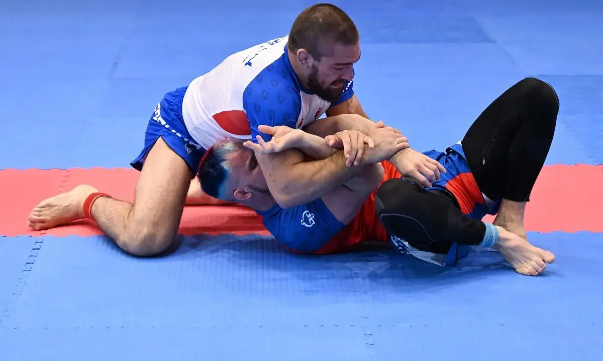 Dwóch mężczyzn walczy w brazylijskim jiu jitsu na niebiesko-czerwonej macie. Jeden z nich klęczy, drugi leży.