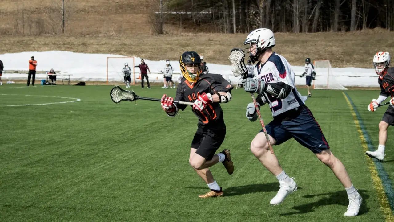 Dwóch zawodników w akcji podczas meczu lacrosse. Jeden biegnie z pałką, drugi go ściga.
