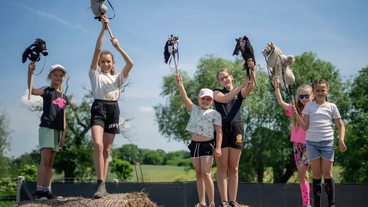 Grupa dziewczynek z dumą prezentuje swoje koniki na kiju, celebrując hobby horsing na tle zieleni.