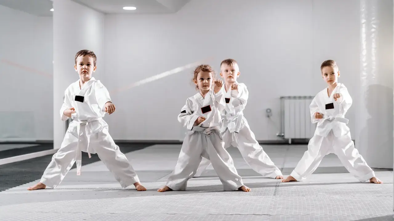 Cztery dzieci w białych kimonach ćwiczą judo dla dzieci. Stoją w pozycji bojowej na macie w jasnej sali treningowej.