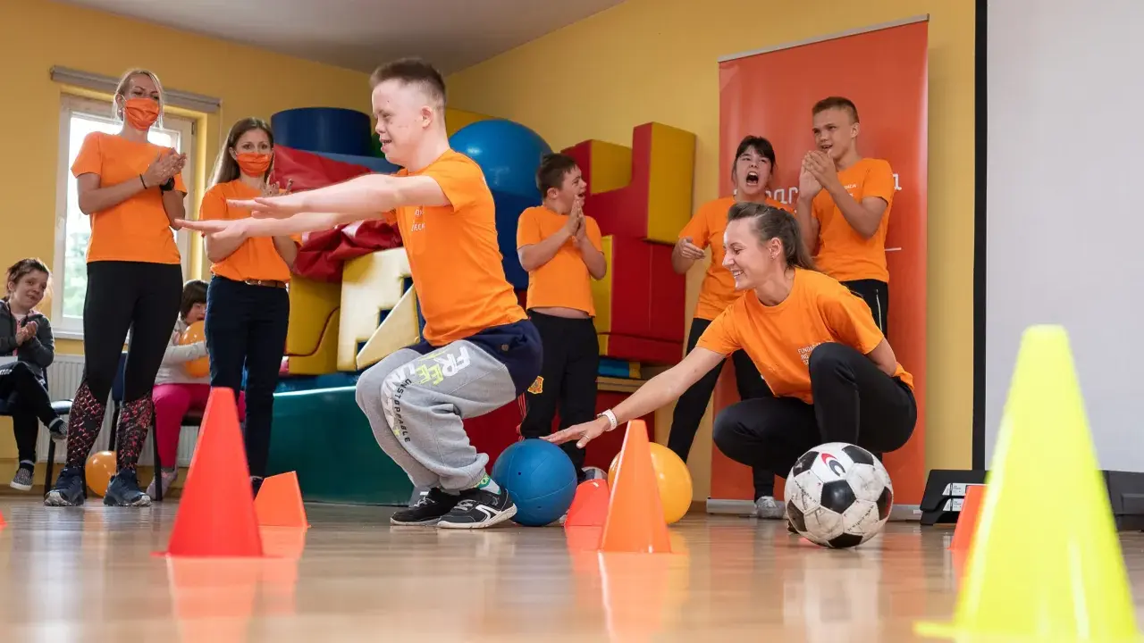 Wesołe wf dla dzieci z piłką i pachołkami. Uśmiechnięci uczestnicy i trenerzy w pomarańczowych koszulkach.