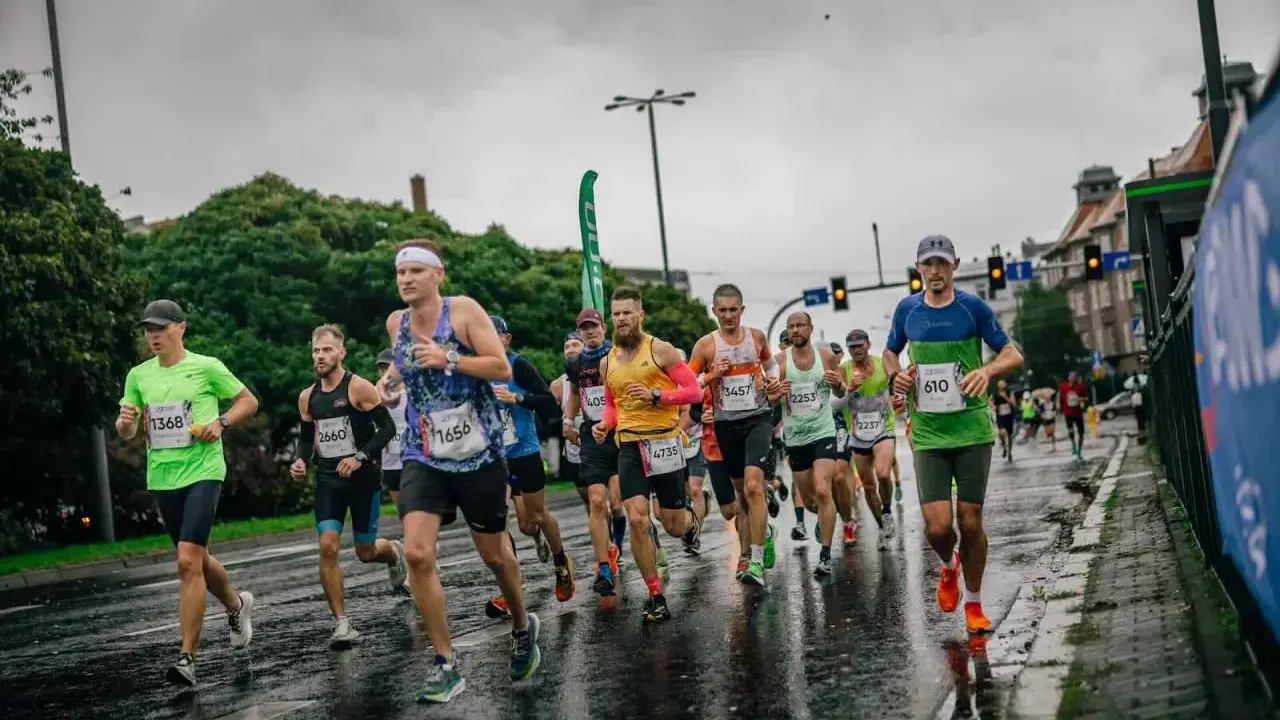 Zawodnicy biegną w deszczu podczas maratonu. Mokra nawierzchnia odbija światła, a biegacze w kolorowych strojach walczą o jak najlepszy czas.