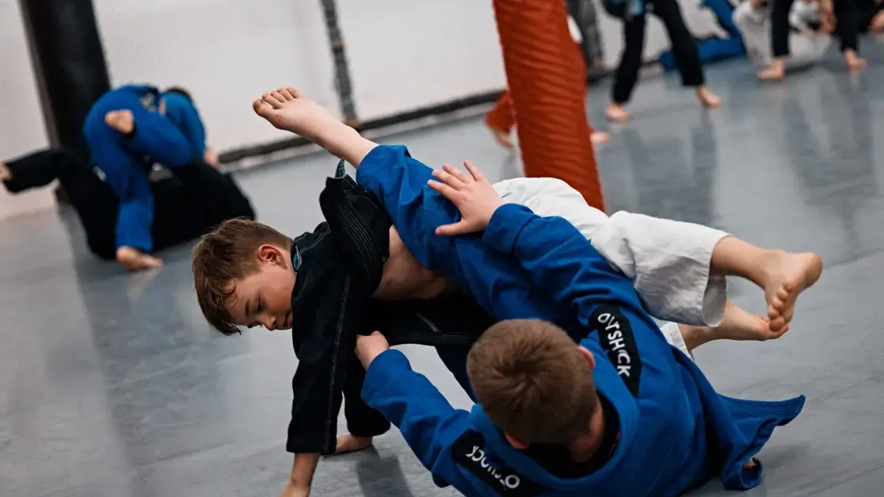 Dzieci ćwiczą jujitsu, walcząc na macie w niebieskich i czarnych kimonach.