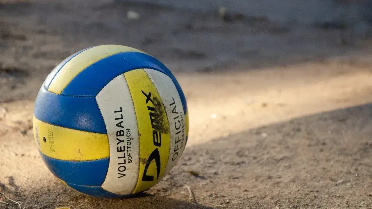 Piłka do siatkówki plażowej, niebiesko-żółta, leży na piasku. Różni się od halowej miękkością i wagą.