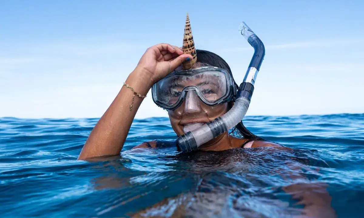 Dziewczyna w masce i fajce do snorkelingu trzyma muszlę na głowie. To właśnie snorkeling!