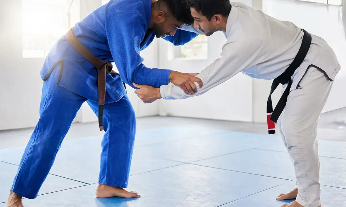 Dwóch mężczyzn w kimonach do BJJ, jeden w niebieskim z brązowym pasem, drugi w białym z czarnym pasem, przygotowuje się do walki.