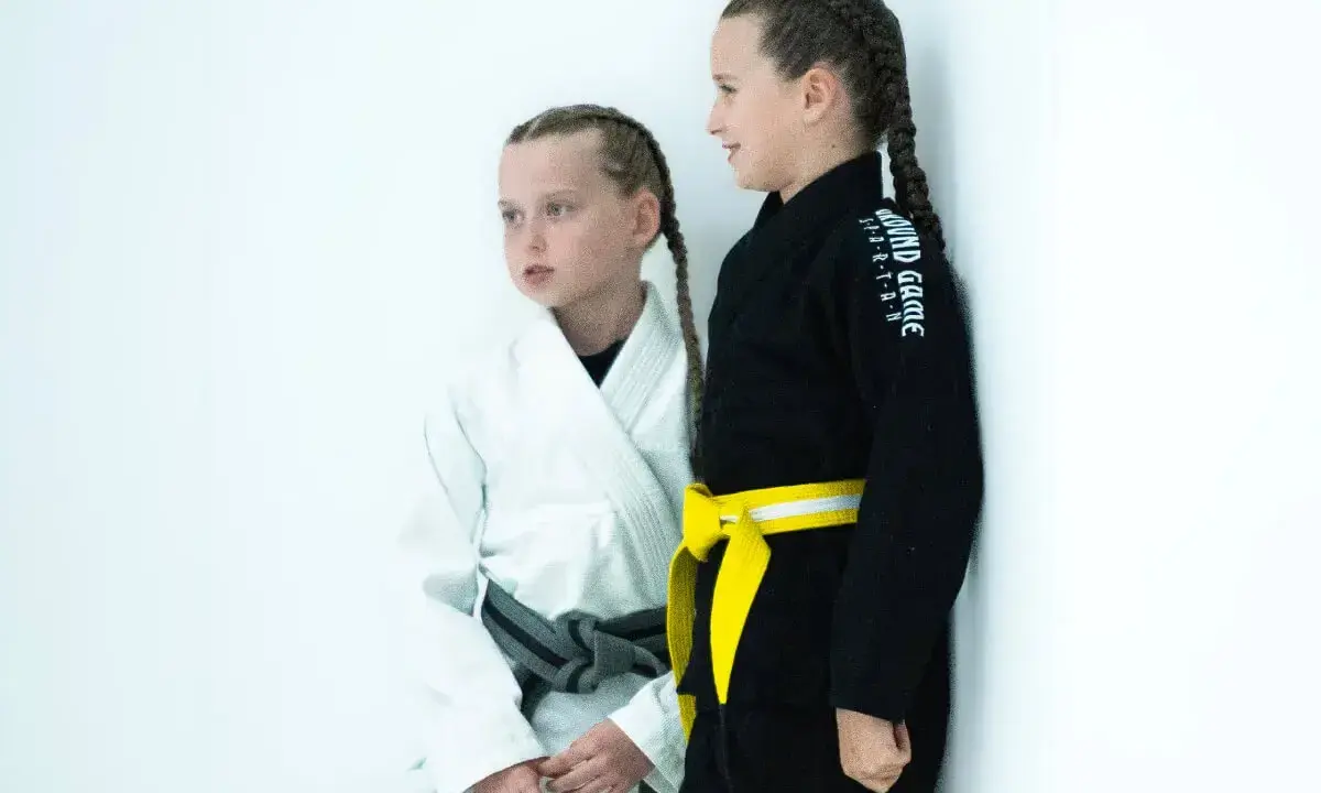 Dwie dziewczynki w kimonach do BJJ, jedna w białym z szarym pasem, druga w czarnym z żółtym pasem. Ich pasy bjj dzieci to symbol postępów w treningach.