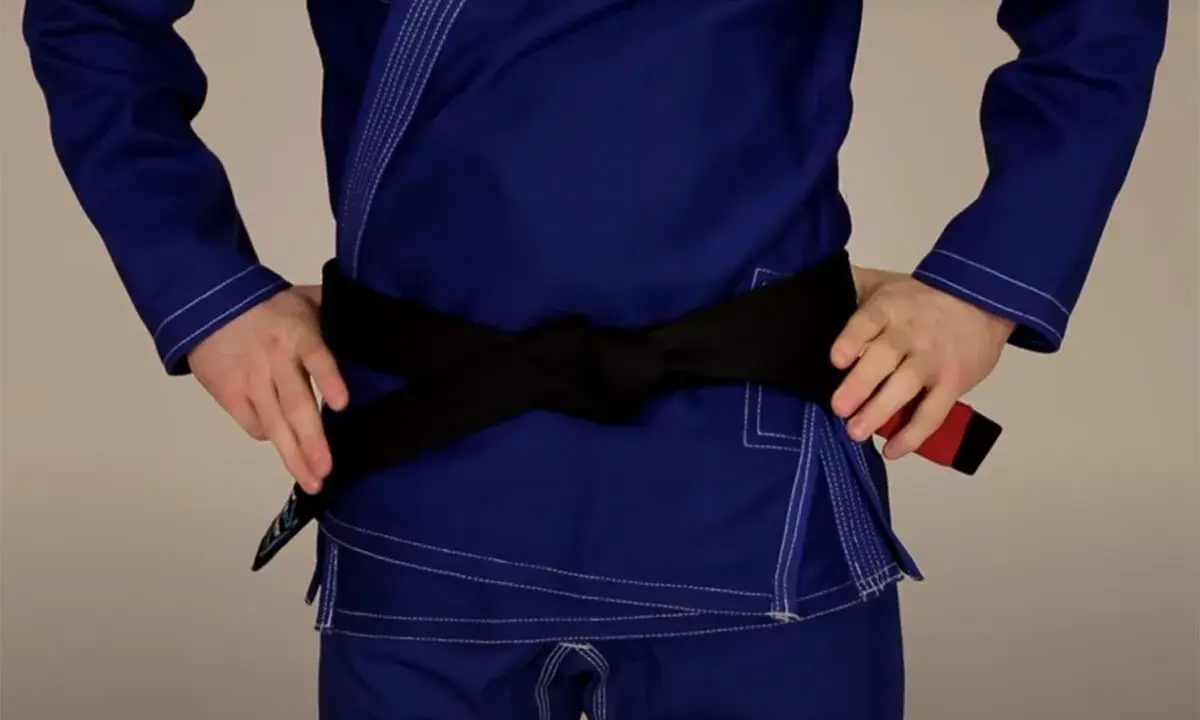 Osoba w niebieskim gi demonstruje wiązanie pasa bjj. Czarny pas jest ciasno zawiązany na wysokości talii.