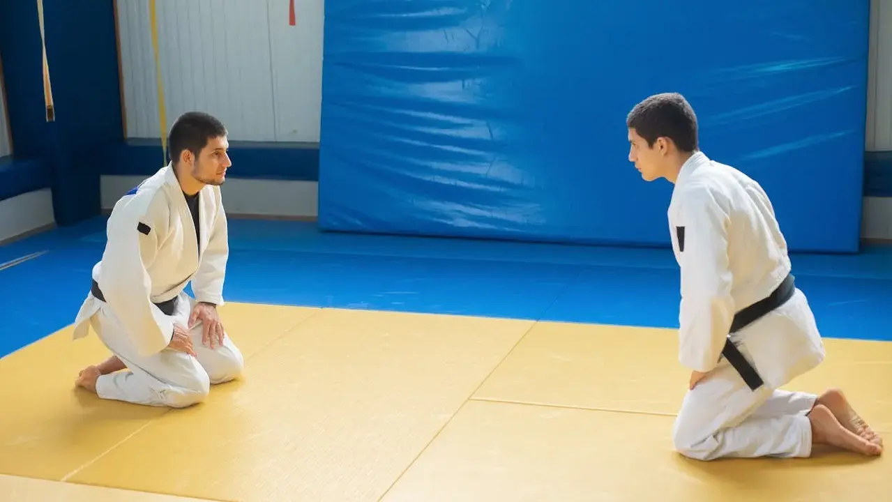 Dwóch judoków w białych kimonach, jeden z czarnym pasem judo, kłania się sobie na macie.