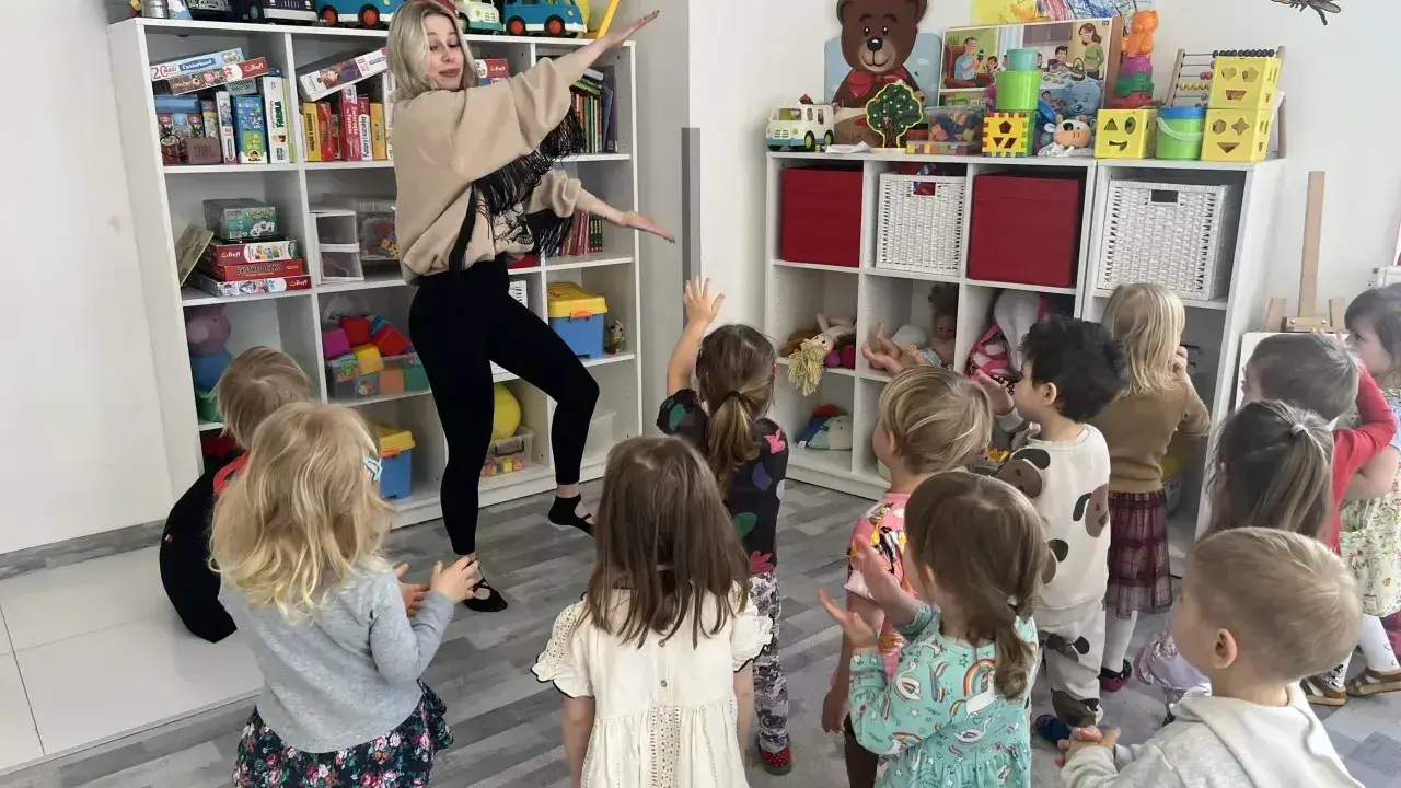 Wesołe zajęcia rytmiczne w przedszkolu. Nauczycielka tańczy z dziećmi wśród zabawek na półkach.