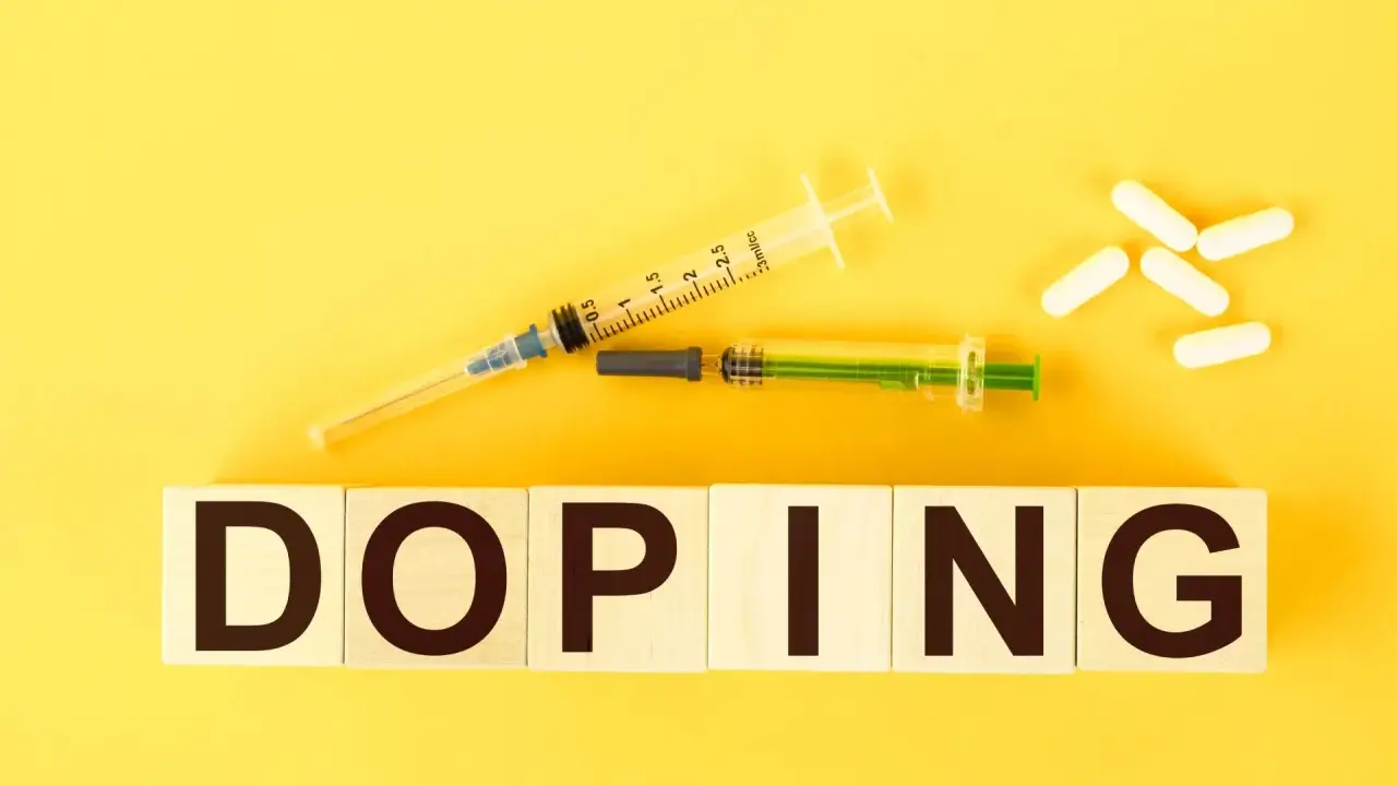 Dwa strzykawki i białe kapsułki na żółtym tle. Słowo "DOPING" napisane na drewnianych klockach.