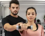 Trening anaerobowy z trenerem. Kobieta ćwiczy z hantlem, mężczyzna koryguje jej ruchy.