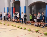 Dzieci w hali sportowej czekają na rozpoczęcie gry i zabawy. Na parkiecie leżą żółte piłki.