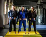 Trzy kobiety w kimonach do BJJ. Środkowa ma purpurowy pas bjj, a pozostałe niebieskie.