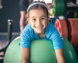 Uśmiechnięta dziewczynka na zielonej piłce gimnastycznej. W tle sztangi, idealne zajęcia sportowe dla 3 latka.