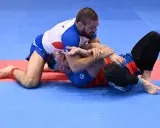 Dwóch mężczyzn walczy w brazylijskim jiu jitsu na niebiesko-czerwonej macie. Jeden z nich klęczy, drugi leży.