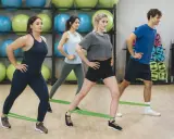 Grupa osób ćwiczy z gumami oporowymi. W tle widać piłki gimnastyczne. To świetny abt trening!