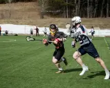 Dwóch zawodników w akcji podczas meczu lacrosse. Jeden biegnie z pałką, drugi go ściga.