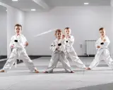 Cztery dzieci w białych kimonach ćwiczą judo dla dzieci. Stoją w pozycji bojowej na macie w jasnej sali treningowej.