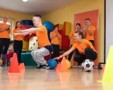 Wesołe wf dla dzieci z piłką i pachołkami. Uśmiechnięci uczestnicy i trenerzy w pomarańczowych koszulkach.