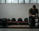 Mężczyzna z tatuażami odpoczywa po treningu crossfit w domu, otoczony piłkami lekarskimi i sztangą.