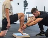 Fizjoterapeuta pomaga starszej osobie w ćwiczeniach równowagi na specjalnej platformie. Konspekt ćwiczeń dla osób starszych.