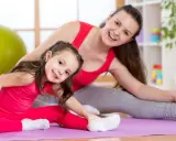 Mama i córka wykonują ćwiczenia rozciągające dla dzieci na macie. Uśmiechnięte, w różowych i szarych strojach.