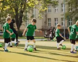Dzieci ćwiczą na boisku w ramach szkółki piłkarskiej. W zielonych strojach biegają z piłkami, ucząc się gry.