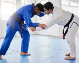 Dwóch mężczyzn w kimonach do BJJ, jeden w niebieskim z brązowym pasem, drugi w białym z czarnym pasem, ćwiczy chwyty.