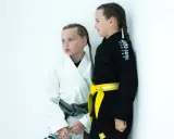 Dwie dziewczynki w kimonach do BJJ, jedna w białym z szarym pasem, druga w czarnym z żółtym pasem. Ich pasy bjj dzieci to symbol postępów w treningach.