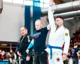Trzech mężczyzn na macie. Jeden w białym kimonie z niebieskim pasem, z podniesioną ręką, ogłoszony zwycięzcą.