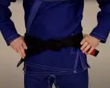 Osoba w niebieskim gi demonstruje wiązanie pasa bjj. Czarny pas jest ciasno zawiązany na wysokości talii.