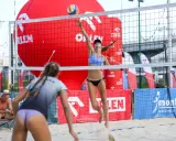 Dwie zawodniczki grają w siatkówkę plażową. Jedna z nich właśnie serwuje piłkę, druga jest w gotowości do obrony.