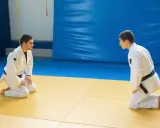 Dwóch judoków w białych kimonach, jeden z czarnym pasem judo, kłania się sobie na macie.