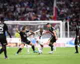 Piłkarze walczą o piłkę, przestrzegając zasad gry. W akcji widać zawodników w białych i czarnych strojach, zmagających się na zielonej murawie stadionu.