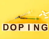 Dwa strzykawki i białe kapsułki na żółtym tle. Słowo "DOPING" napisane na drewnianych klockach.