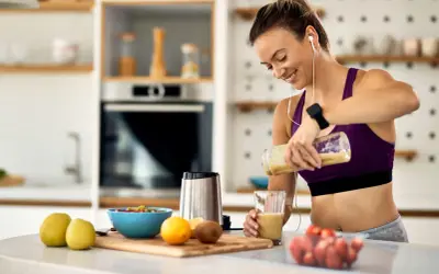 Uśmiechnięta kobieta przygotowuje smoothie, idealne jako co zjesc przed biegiem. Na blacie owoce i blender.