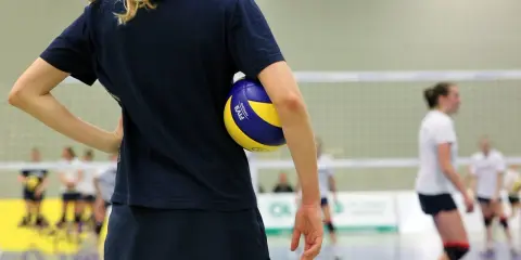 Dziewczyna w granatowej koszulce i spódniczce trzyma piłkę do siatkówki. Na drugim planie inne zawodniczki.