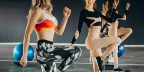Dwie kobiety ćwiczą na stepach. Jedna w kolorowym topie i wzorzystych legginsach, druga w czarnym body i beżowych legginsach. TBC co to znaczy?