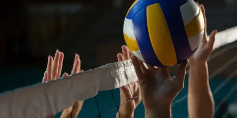 Piłka do siatkówki tuż nad siatką, gracze próbują ją odbić. Widać ich dłonie i fragmenty siatki, która wyznacza wysokość siatki w siatkówce.