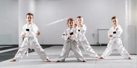 Cztery dzieci w białych kimonach ćwiczą judo dla dzieci. Stoją w pozycji bojowej na macie w jasnej sali treningowej.
