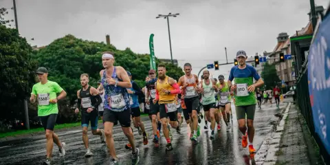 Zawodnicy biegną w deszczu podczas maratonu. Mokra nawierzchnia odbija światła, a biegacze w kolorowych strojach walczą o jak najlepszy czas.