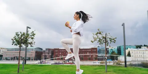 Kobieta w białym stroju sportowym wykonuje dynamiczne ćwiczenie w parku. W tle miejska panorama.