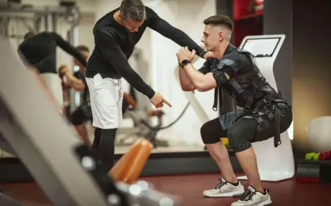 Trening EMS: Instruktor koryguje pozycję mężczyzny w kamizelce stymulującej mięśnie podczas przysiadu.