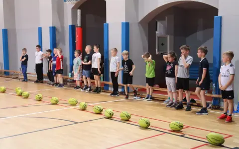 Dzieci w hali sportowej czekają na rozpoczęcie gry i zabawy. Na parkiecie leżą żółte piłki.